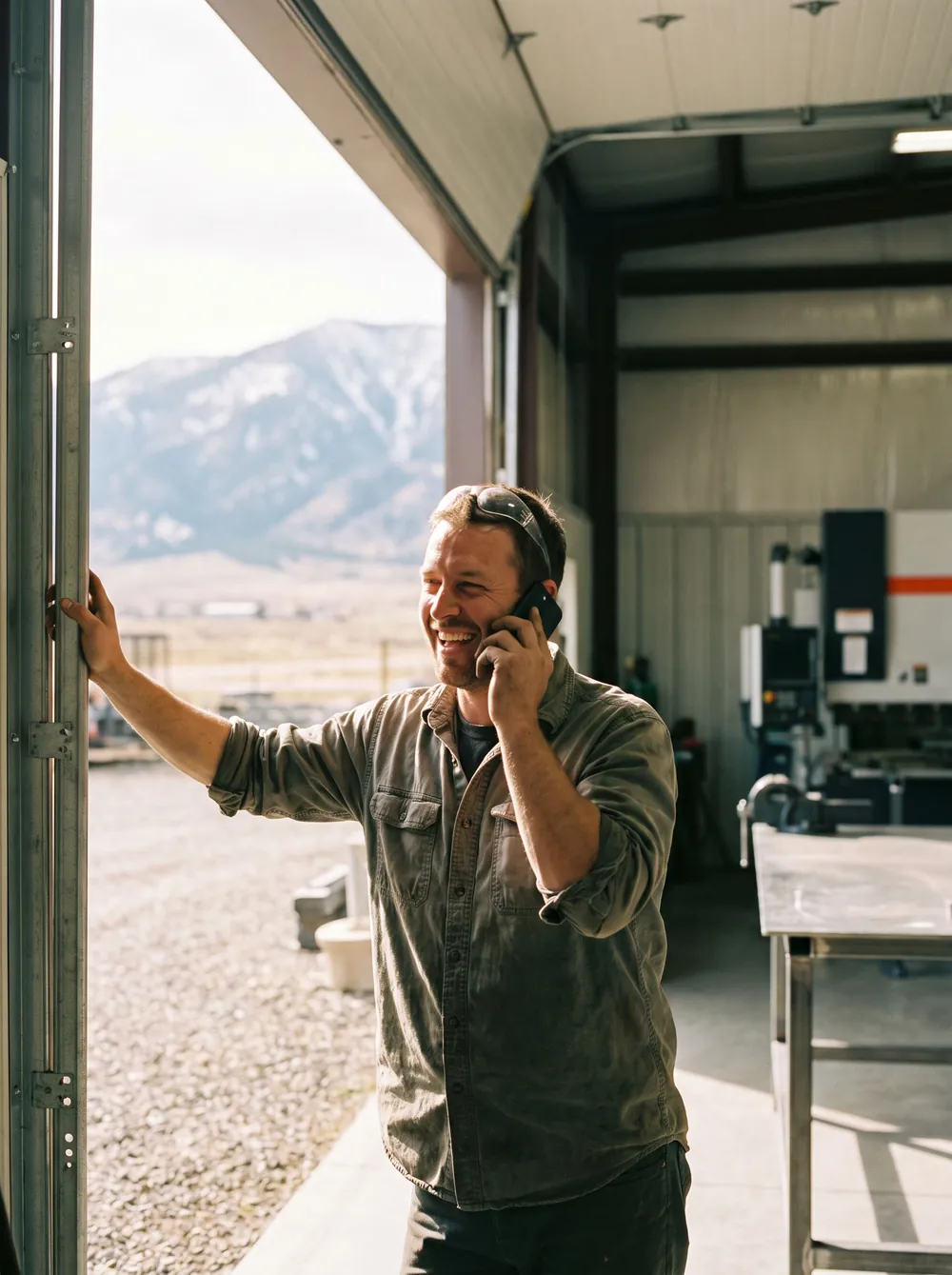 A metal fabricator laughing on a phone call in the open door of his shop, a snow-dusted range behind him